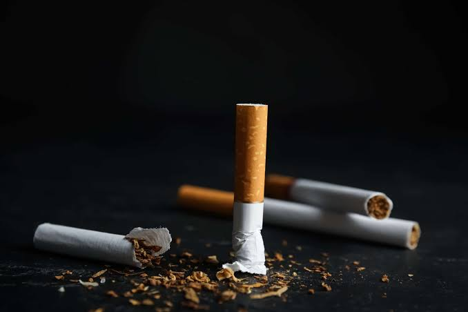 A broken tobacco cigarette standing upright next to loose dried tobacco shreds and unlit filter cigarettes on a dark reflective surface - Nicotine in a Cigarette vs Vape comparison by VapeFeat
