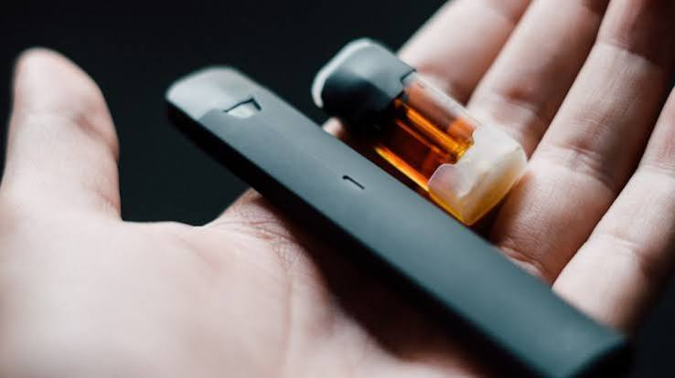 Close-up of an open hand holding a sleek black pod vape battery alongside a detached clear e-liquid pod cartridge filled with golden vape juice - Vapes May Help Smokers Quit guide by VapeFeat