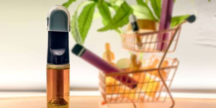 Amber cannabis oil vape cartridge standing in front of miniature rose gold shopping carts filled with vape pens and botanical wellness products - Anxiety Pen vs Vape guide by VapeFeat
