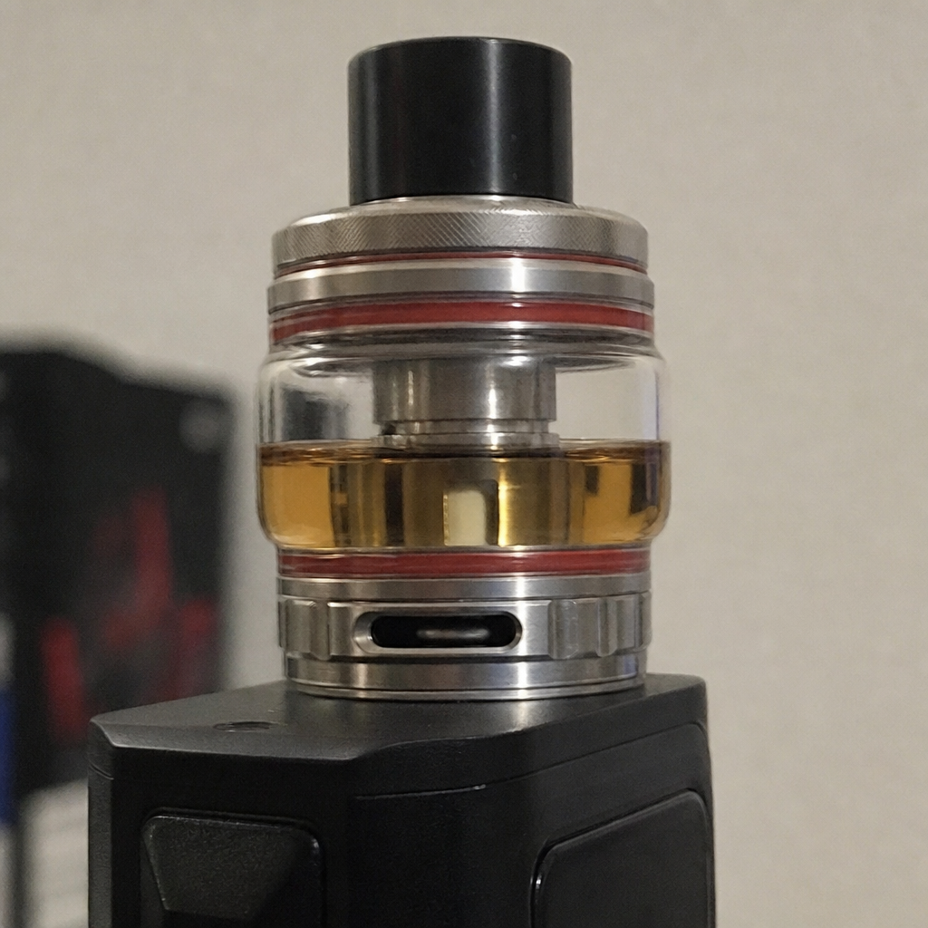 Close-up of a stainless steel sub-ohm vape tank filled with golden e-liquid, featuring red O-rings, bubble glass, and bottom airflow slot - Vape Tanks guide by VapeFeat