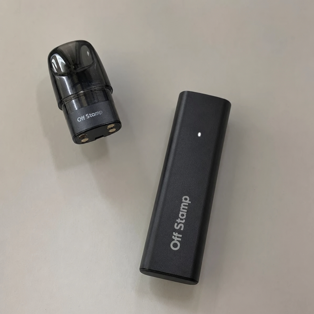 Black Off Stamp rechargeable magnetic battery dock with lit LED indicator beside a detached prefilled disposable pod cartridge showing contact pins on a light surface - Off Stamp Vape review by VapeFeat