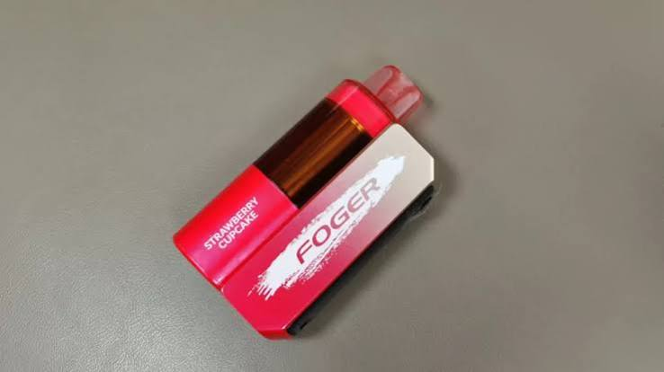 Foger Switch disposable vape in Strawberry Cupcake flavor lying at an angle on a grey surface showing visible transparent tank and silver battery casing - Foger Vape review by VapeFeat