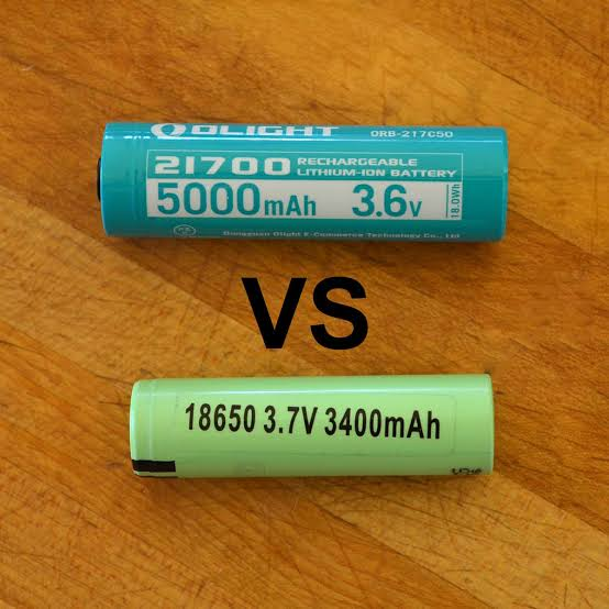 Blue Olight 21700 5000mAh battery compared against a green 18650 3400mAh cell on a wooden surface - 18650 vs 21700 vape battery guide on VapeFeat