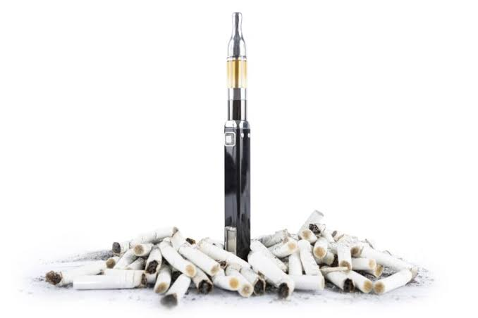 Sleek black e-cigarette vape pen with clear golden tank standing upright inside a mound of discarded cigarette butts on a white background - Vaping vs Smoking Harm Reduction Guide on VapeFeat