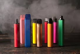 Variety of colorful disposable vape pens and bar devices standing upright on a rustic dark wooden surface against a smoky backdrop - Best Disposable Vapes Guide on VapeFeat