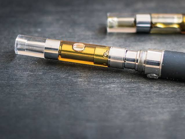 Close-up of a 510 thread glass vape cartridge filled with golden distillate cannabis oil attached to a black battery pen on a dark slate background - VapeFeat