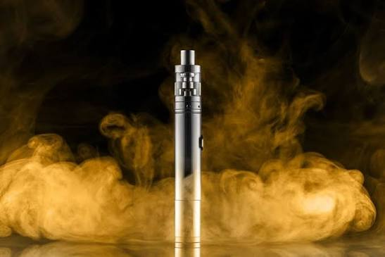 Stainless steel vape pen tube mod standing upright on a reflective surface surrounded by dramatic golden vapor smoke clouds