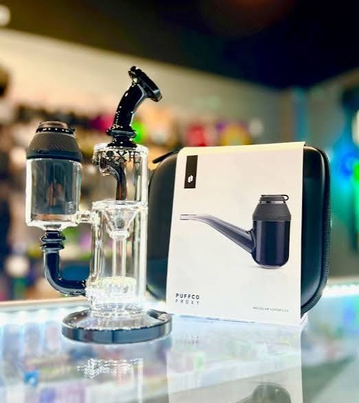 Puffco Proxy modular vaporizer base slotted into a clear percolator glass bubbler attachment next to original travel case packaging on a dispensary counter