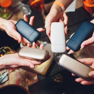 Group of people holding colorful multi-generation IQOS heat-not-burn (HNB) device pocket chargers and holders together over a social gathering table - VapeFeat